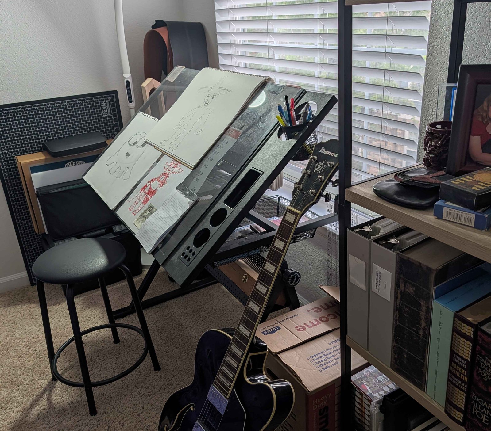 A corner workspace: a drafting table with sketches pinned to the board, a stool, a hollow-body guitar leaning against it, shelves of books and boxes, lit by a window